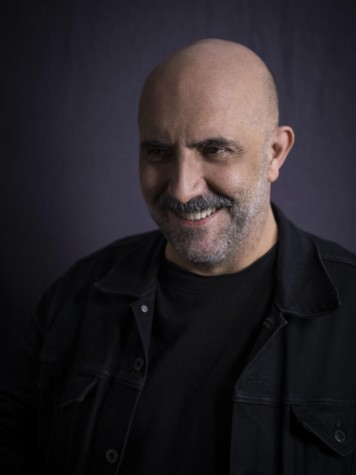 A portrait of a smiling Gaspar Noé wearing all black in front of a gray background.