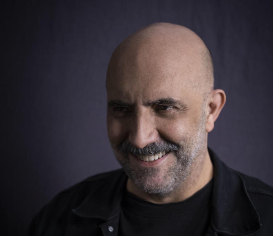 A portrait of a smiling Gaspar Noé wearing all black in front of a gray background.