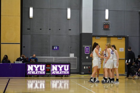 At a basketball court with posters that read “NYU Athletics,” the NYU Women’s Basketball team are gathered in a circle.