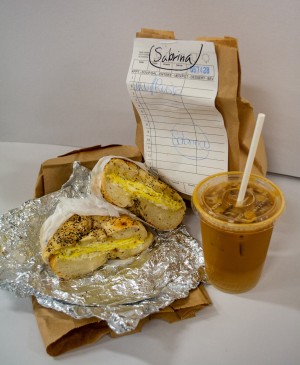 A picture of a scrambled eggs and cream cheese bagel cut in half against a white background. Behind it a brown bag with a receipt that reads “Sabrina.” Next to the bagel is a plastic cup of coffee.