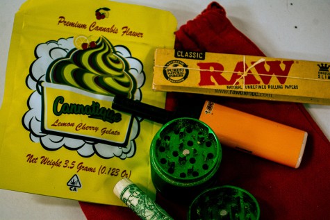 A yellow bag, rolling papers, an orange lighter, a dollar bill, and a small green grinder on top of a red bag.