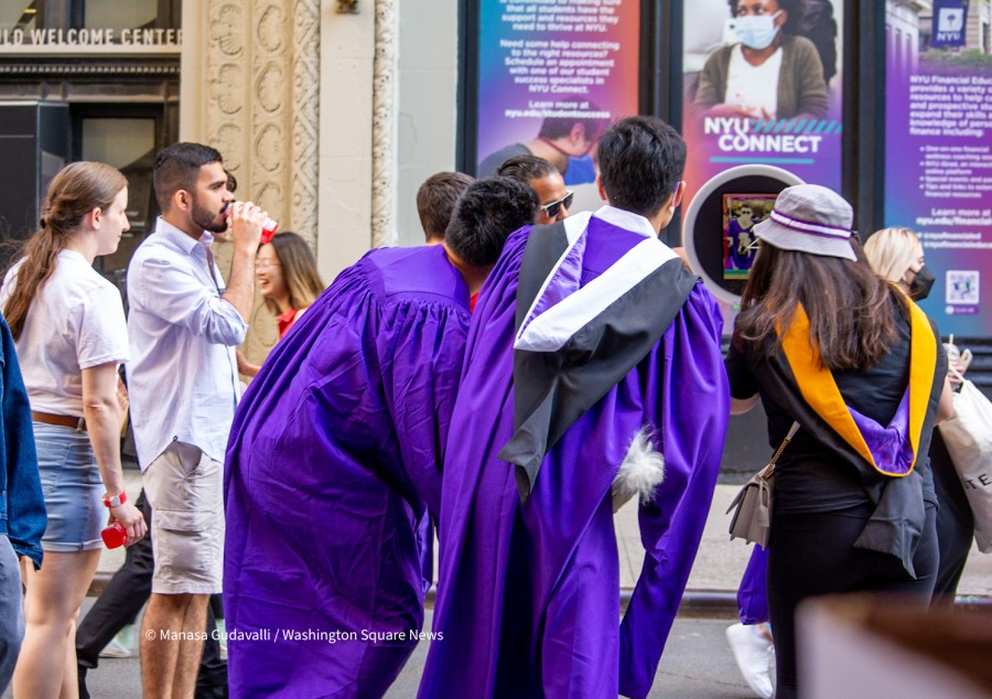 Two graduates in purple graduation robes crouch in front of a ring light camera to take a picture. There are other graduates, friends and family members walking around them. In the background, there banners for NYU Connect.
