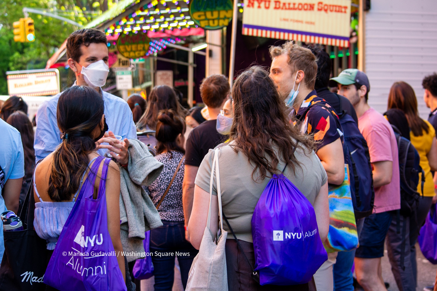 A group of graduates stand on the street with purple bags with the white text reading “NYU Alumni Bags.” Behind them are crowds of people and carnival tents.