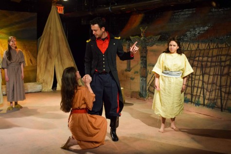 A man wearing a red and black soldier uniform stands with his right hand holding the hand of a woman on her knees wearing a long orange dress. The man is raising his left hand in a speaking motion. They stand on a theater stage. To the right, there is a woman wearing a long yellow dress.