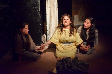 A woman in a long yellow dress sits on her knees, illuminated by a spotlight. Behind her there is a white column, and to both of her sides there are women in long gray dresses holding her hands.