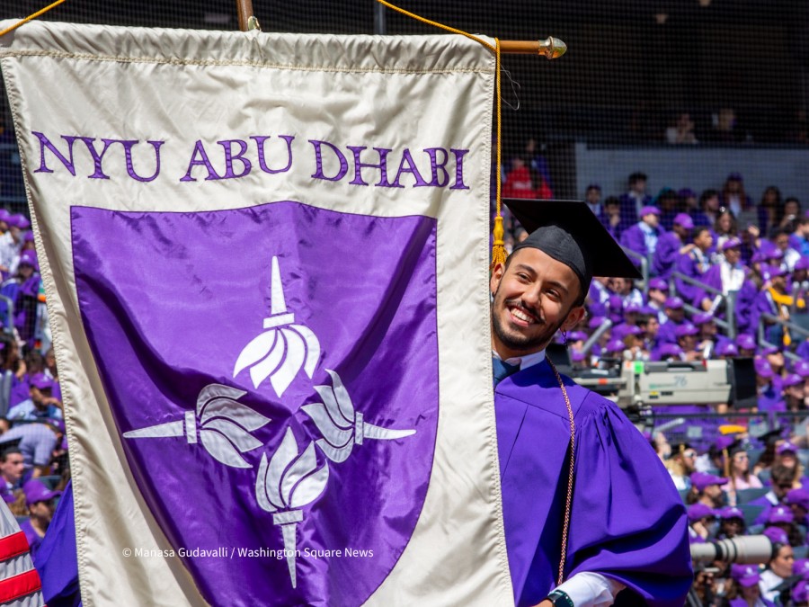 Graduate+Ayham+Adawi+in+purple+graduation+robe+holds+the+NYU+Abu+Dhabi+banner.+A+crowd+of+graduates+in+purple+robes+are+seen+in+the+background.