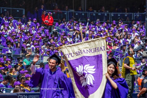 Graduates Matthew Fertig, left, and Mengjie Shen, right, in purple graduation robes hold the NYU Shanghai banner. A crowd of graduates in purple robes are seen in the background.