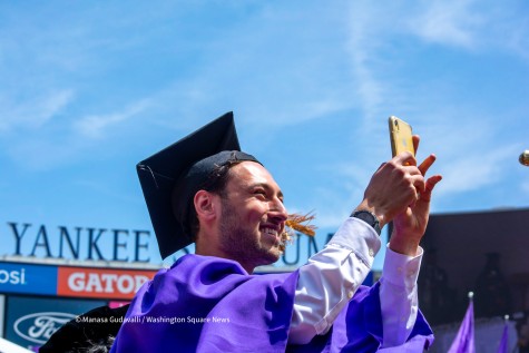 NYU graduate Matthew Fertig in a purple graduation robe holds a phone in front of him. The Yankee Stadium sign is seen in the background.