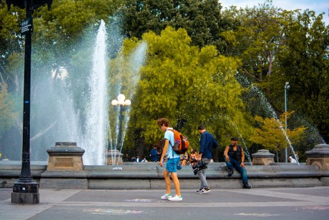 A student walks past the fountain in Washington Square Park. Behind the fountain are green trees with lots of foliage. It is a sunny day with a bright blue sky full of clouds. The fountain in the center of the park is on.