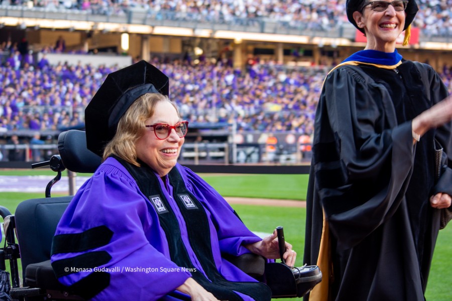 Judith Heumann, wearing violet academic robes and red spectacles, enters Yankee Stadium during NYUs commencement ceremony for the classes of 2020 and 2021. She smiles and controls her wheelchair with one hand. Faye Ginsburg is on her left, wearing a black robe and smiling. A crowd of graduates are in the stands in the background.