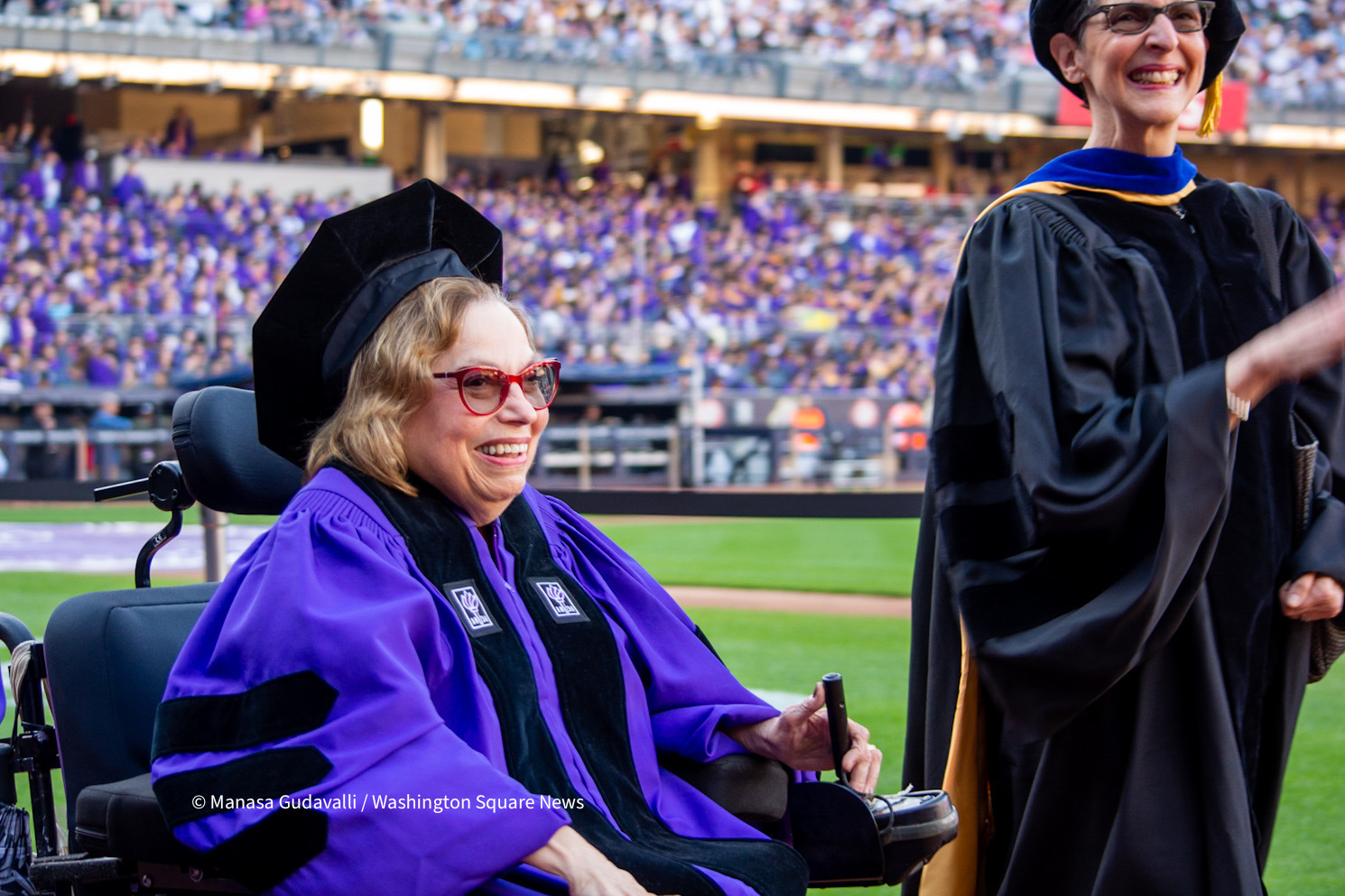 Judith Heumann, wearing violet academic robes and red spectacles, enters Yankee Stadium during NYU's commencement ceremony for the classes of 2020 and 2021. She smiles and controls her wheelchair with one hand. Faye Ginsburg is on her left, wearing a black robe and smiling. A crowd of graduates are in the stands in the background.