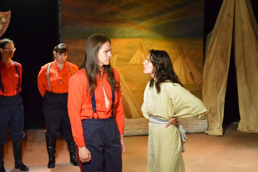 On a theater stage, there is a girl wearing a yellow dress looking toward and talking to a guy wearing a red shirt and blue pants. Behind them is a backdrop with tepees.