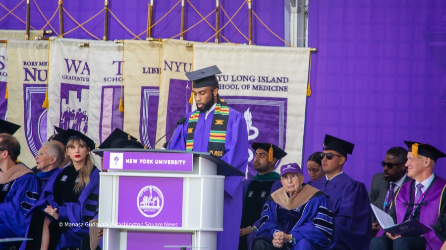 NYU graduate Rodney Anderson in purple graduation robe and Kente stole speaks behind the podium. People sit behind them. NYU president Andrew Hamilton and Taylor Swift look at them. School flags and a purple curtain are in the background.