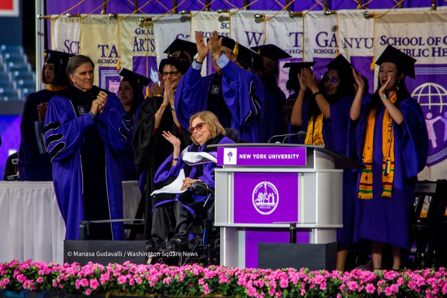 Judith Heumann waves to the crowd while on stage after delivering remarks during NYUs commencement ceremony for the classes of 2020 and 2021. NYU graduates and administrators cheer and clap their hands behind her. The backdrop of the stage is several NYU school flags.