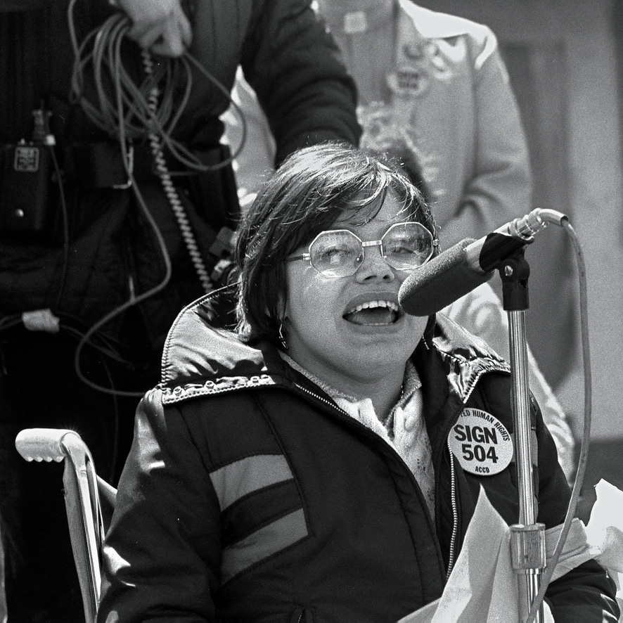 A black-and-white picture of Judith Heumann speaking in front of a microphone. On her jacket, there is a pin that reads “Sign 504.”