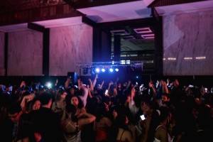 The Bobst atrium at night: formally dressed ball attendee dancing in the foreground; a DJ stand blasting purple light beams in the background.