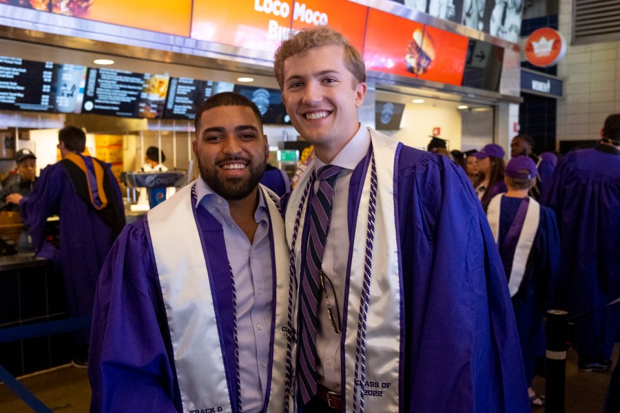 Two NYU graduates, Michael Richards and Michael Manuel Jr., are wearing purple graduation robes and are standing in the food court at Yankee Stadium.