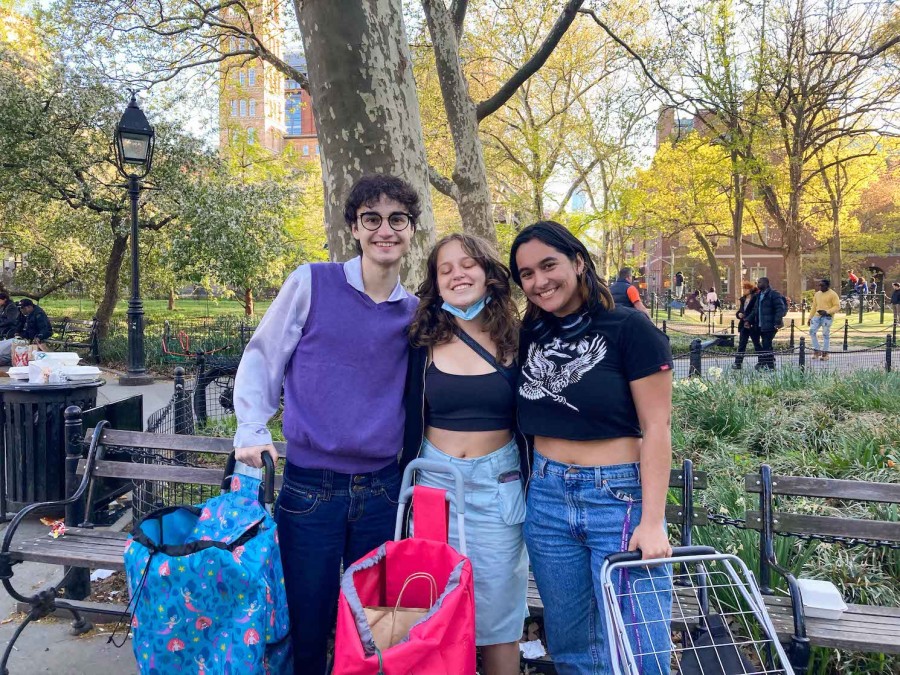 A boy and two girls stand side by side smiling at the camera. Each holds different styles of food shopping bags. Behind them, the green trees of Washington Square Park.