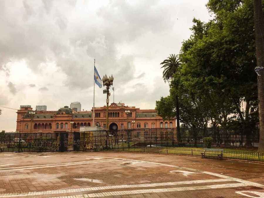 The pink facade of la Casa Rosada, the office of the Argentinian president. In front of it there is a blue-and-white Argentinian flag and a plaza. To the right are green trees.