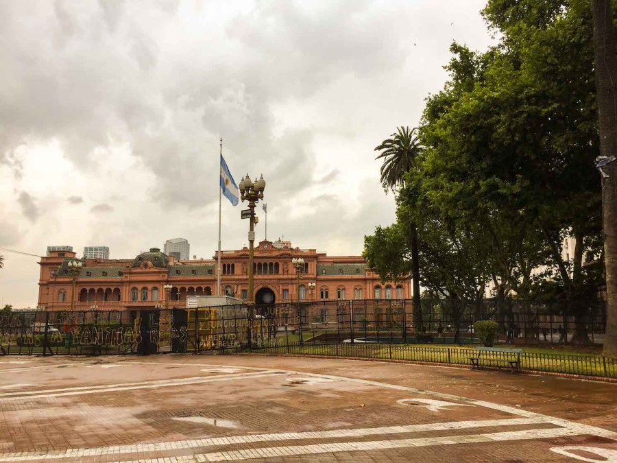 The+pink+facade+of+la+Casa+Rosada%2C+the+office+of+the+Argentinian+president.+In+front+of+it+there+is+a+blue-and-white+Argentinian+flag+and+a+plaza.+To+the+right+are+green+trees.