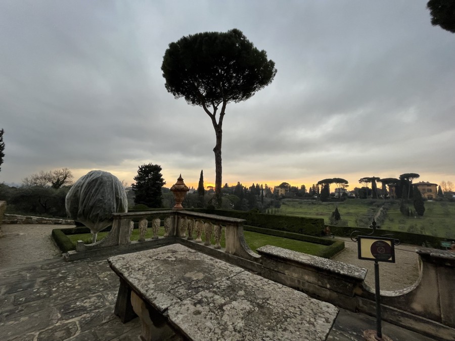 A view of a sunset from a stone patio at NYU Florence, looking across the valley toward Villa Natalia. Trimmed hedges line the path down from the patio and cypress trees adorn the skyline.