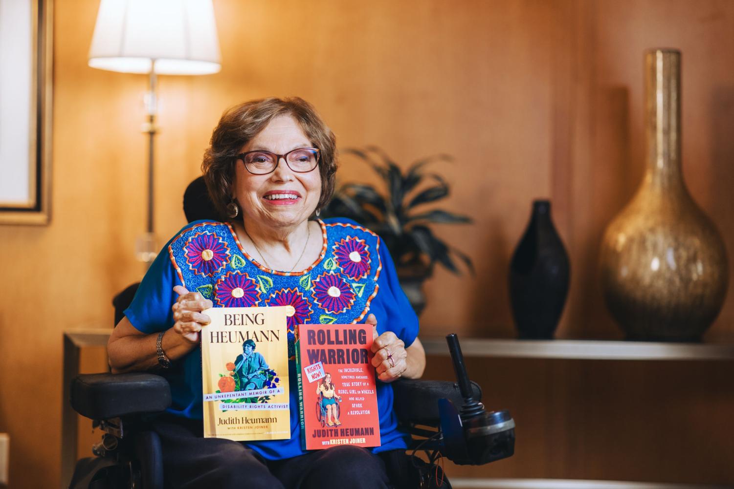 Judith Heumann sits in a warmly lit room holding up two of her books, "Being Heumann" and "Rolling Warrior." She is wearing a blue top with floral embroidery. In the background, two vases sit on a shelf and a lamp can be seen.