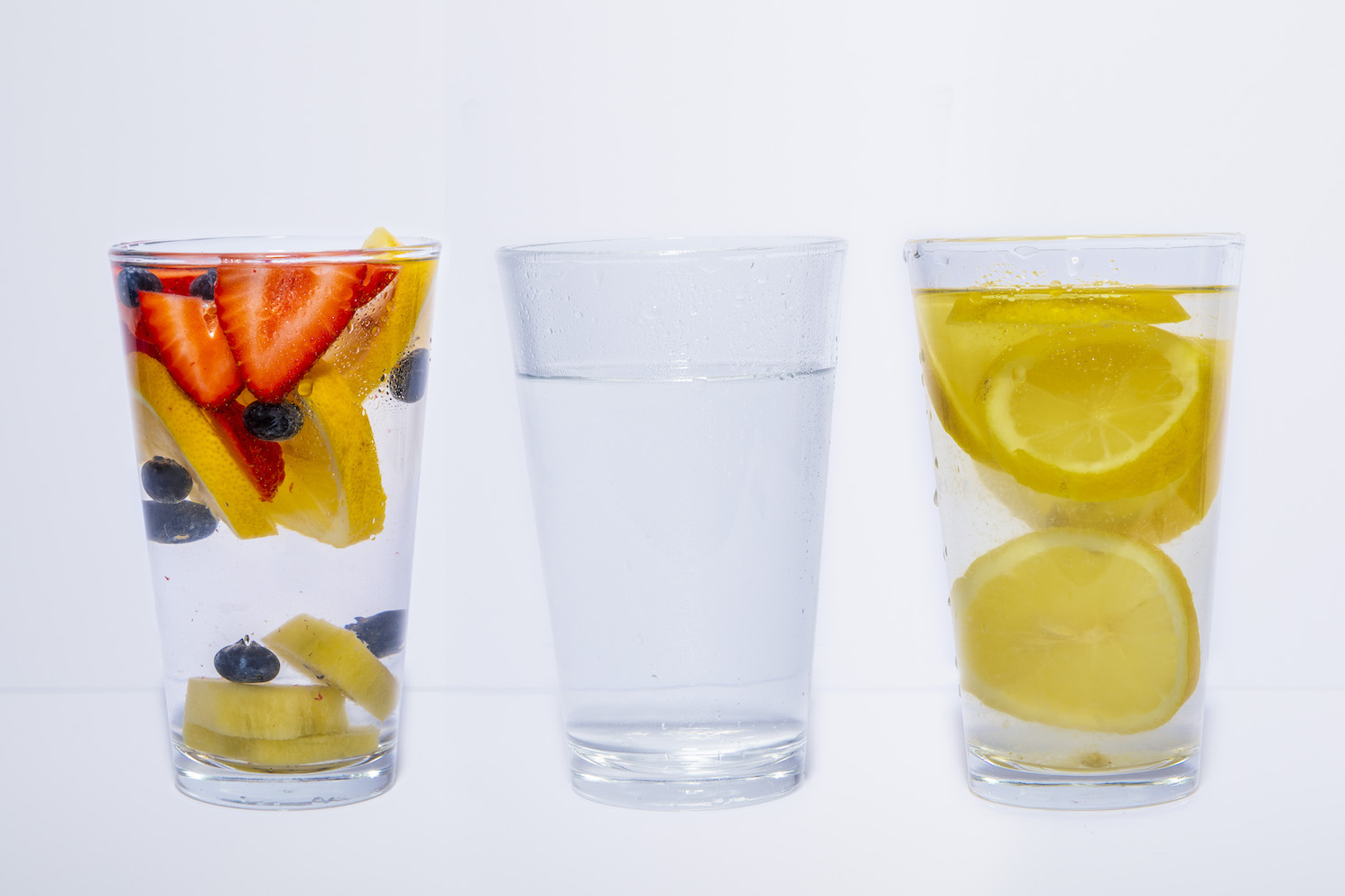Three glasses of water against a white background. Two of the three glasses of water have lemon slices in them, and one of the three also has berries.