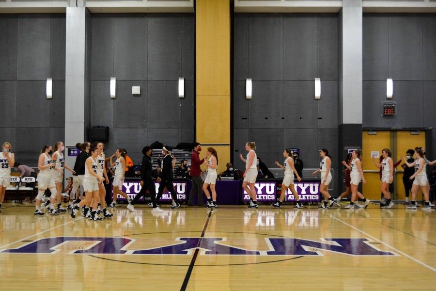 On the NYU basketball court, the NYU Women’s Basketball team stands in a line. Players are clapping the other basketball team’s hands as they walk in the opposite direction.