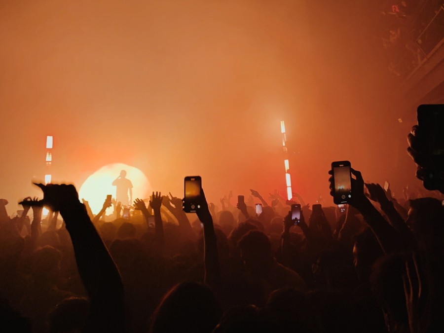 A group of concert attendees holding their phones up high in the foreground, and a performer clouded by orange mist in the background.