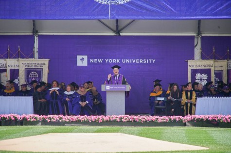 A stage with a purple backdrop and banners representing NYU's schools and colleges hang in the background. A podium is at the center, and NYU president Andrew Hamilton stands behind it wearing a purple academic robe. He is speaking with one hand raised in the air. Surrounding him are seated commencement guests, also dressed in purple academic robes.