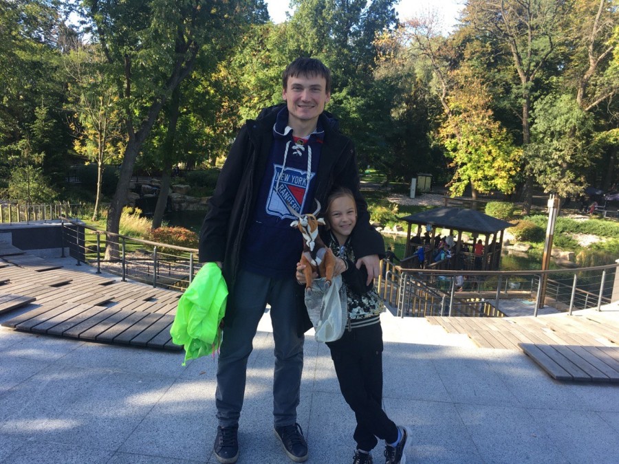 Yuriy Fedchyshyn and his daughter standing next to each other on a pavilion with trees and green foliage in the background. Yuriy has his arm around his daughter, who is holding a stuffed animal.