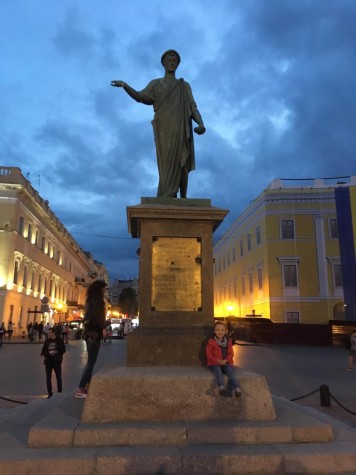 The Monument to Duc de Richelieu in the evening. A young girl is sitting on the steps of the monument. The sun has set and there are buildings with yellow lighting in the background.