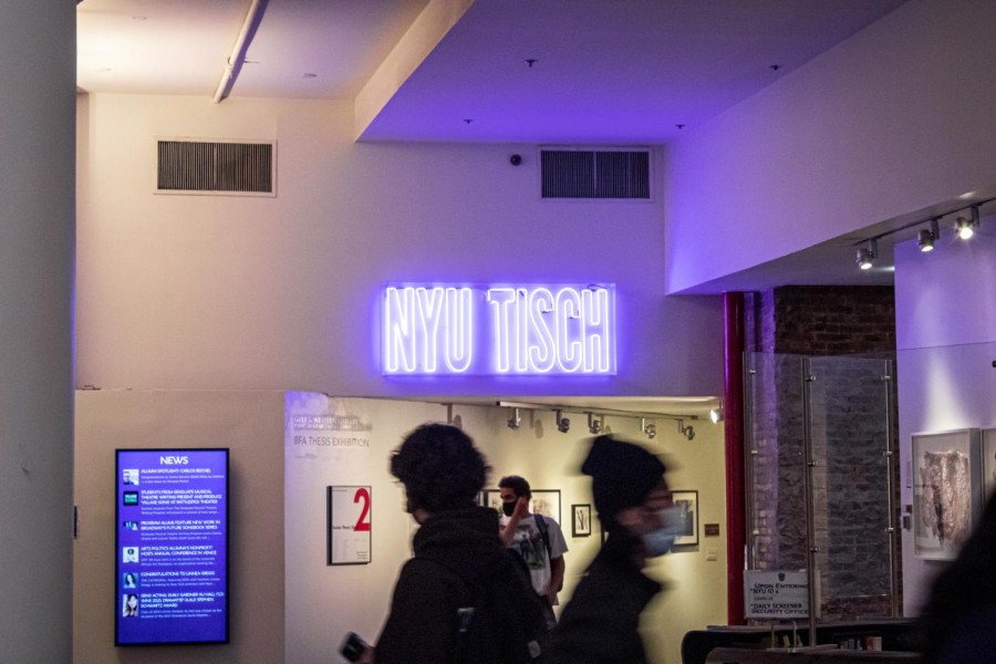 The interior of the NYU Tisch building. “NYU TISCH” is displayed on the wall in a purple LED light sign. People are walking by.