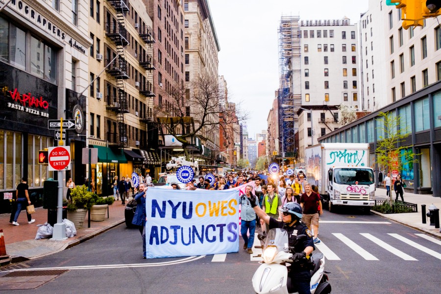 Protesters+walk+down+University+Place+holding+a+large+sign+that+reads+%E2%80%9CNYU+owes+adjuncts.%E2%80%9D+A+police+officer+on+a+moped+directs+traffic+in+front+of+the+marching+protesters.