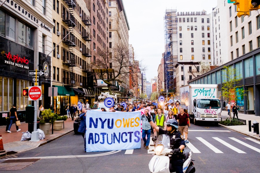 Rally+attendees+walk+down+University+Place+holding+a+large+sign+that+reads+%E2%80%9CNYU+owes+adjuncts.%E2%80%9D+A+police+officer+on+a+moped+directs+traffic+in+front+of+the+marching+protesters.