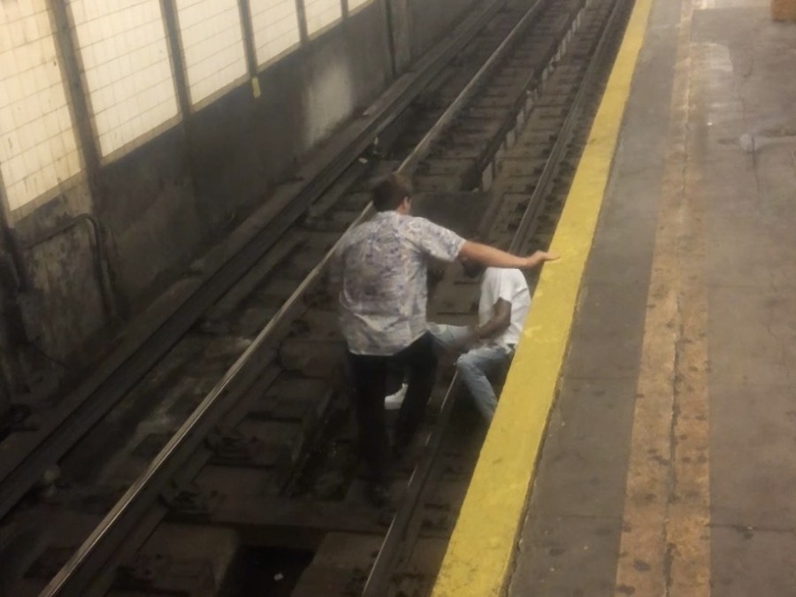 A young man wearing black pants and a light-colored short sleeve button-up shirt holds onto the side of a subway platform, standing on the tracks over another man who is laying down on the tracks.
