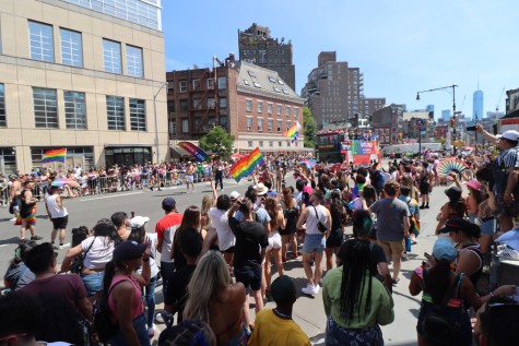 Crowds gather on the sidewalk behind barriers watching as a bus and people pass by in The NYC Pride March.