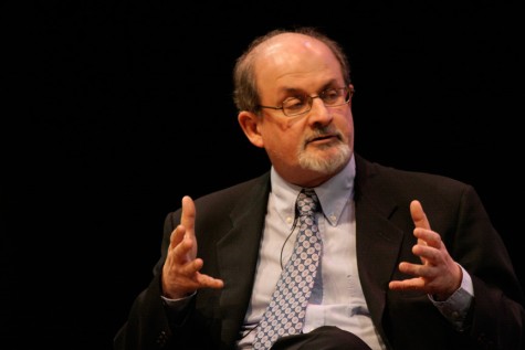 An upper body portrait of Salman Rushdie in a black suit.