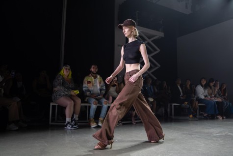A model wearing a black crop top and brown jeans walking down a runway with the audience sitting in the back.