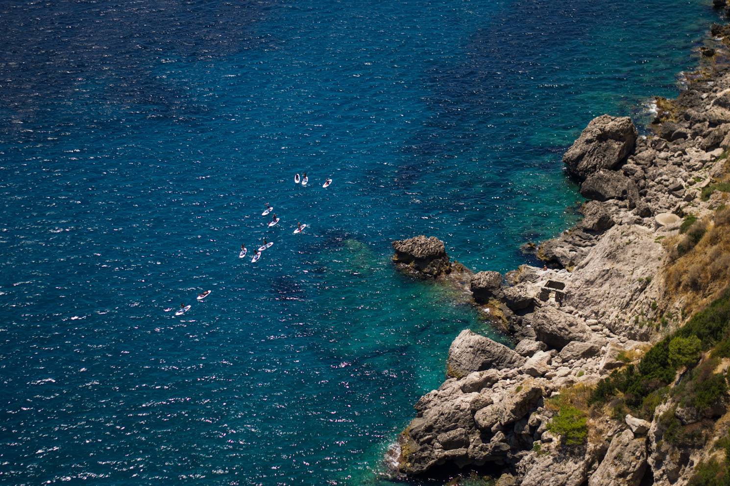 A bird’s-eye view of a light blue ocean with a rocky coastline.