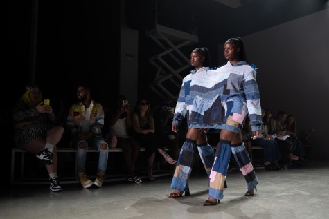 Two models sharing a denim shirt with patches walking down a runway with the audience sitting in the back.