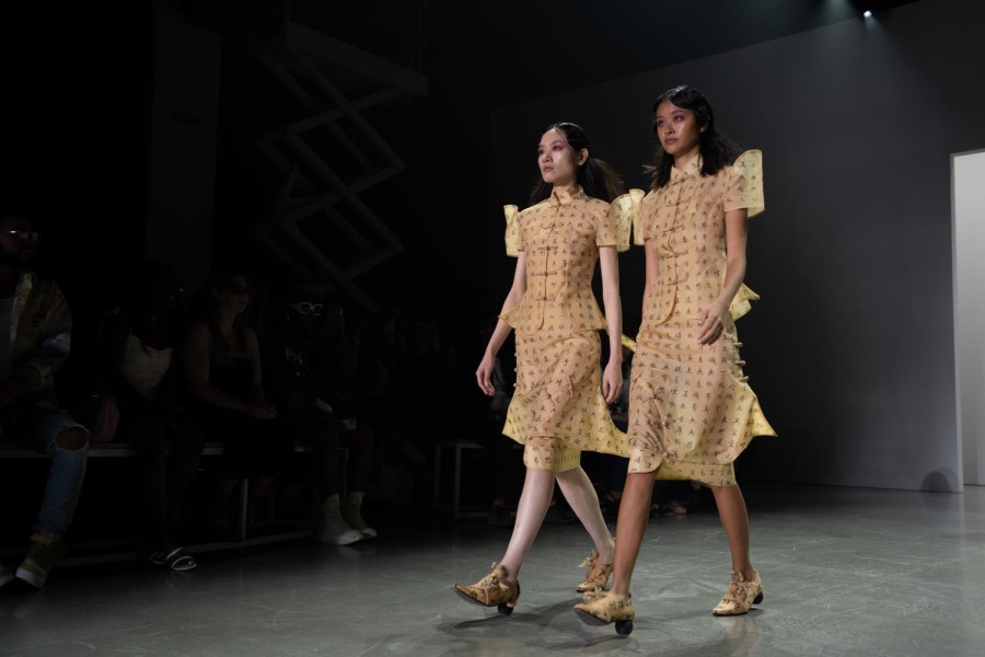 Two models wearing similar yellow dresses with Chinese characters written across them walking side by side down a runway with the audience sitting in the back.