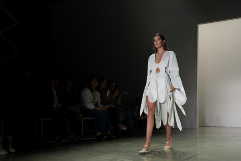 A model wearing a light blue silky blouse and shorts walking down a runway with the audience sitting in the back.