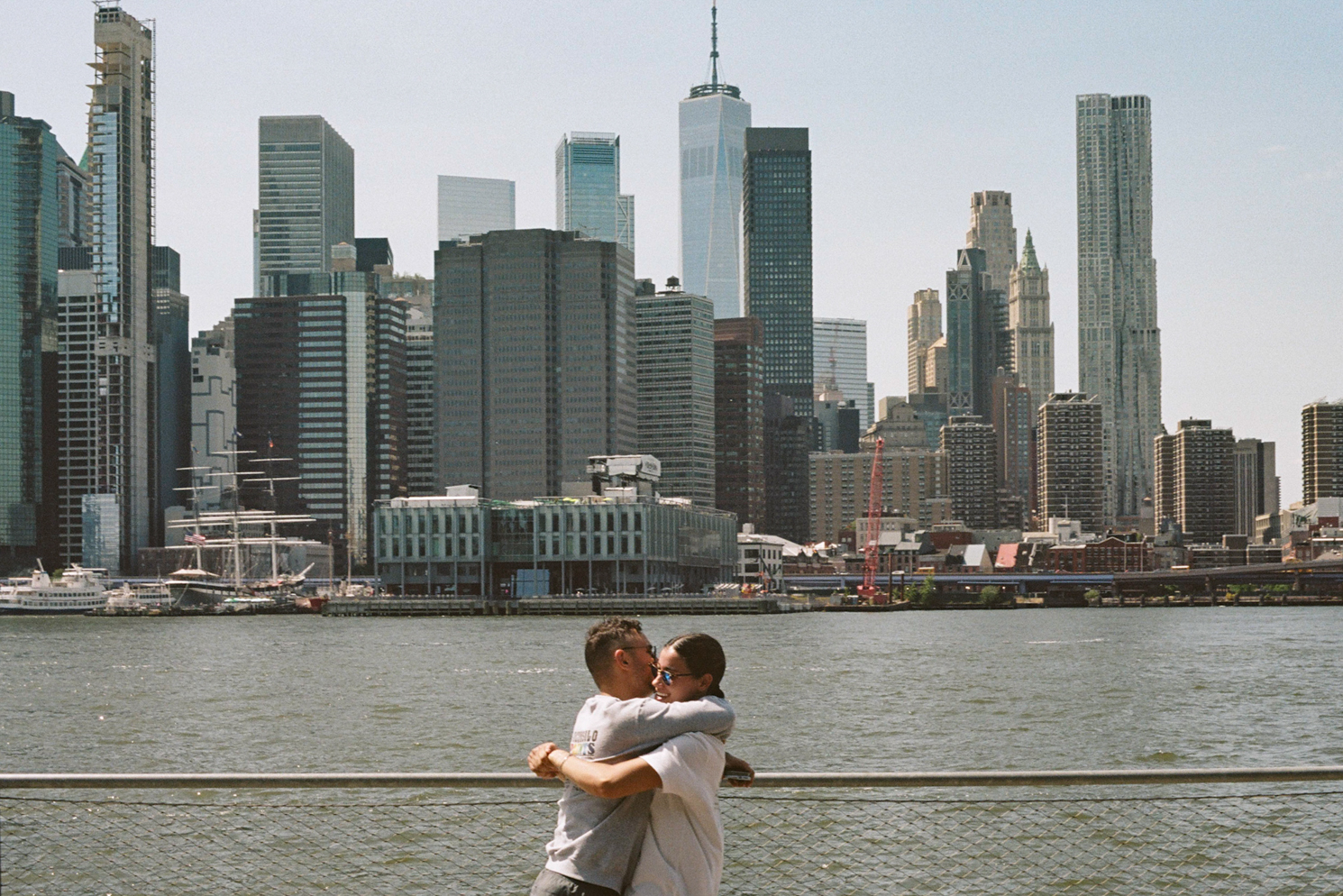 A man and a woman embracing at a waterfront. The downtown Manhattan skyline is in the background.