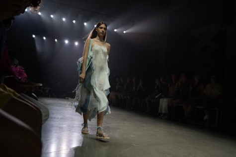 A model wearing a light blue dress and Rome sandals walking down a runway with the audience sitting in the back.