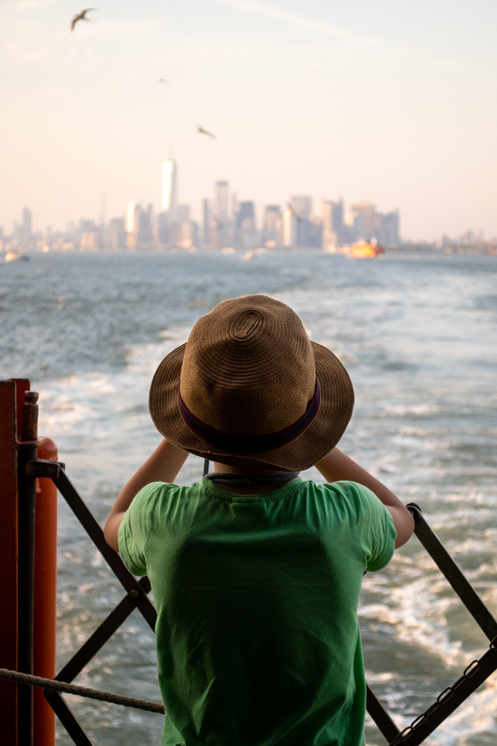 A child wearing a green shirt and a brown hat looking at the Manhattan skyline from the back of a ferry.