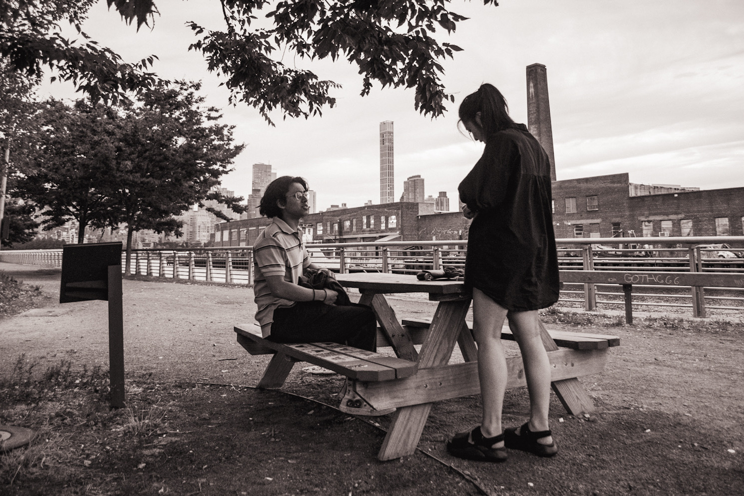 Black-and-white photo of a man sitting at a picnic table with a woman standing next to him. The city skyline is in the background.