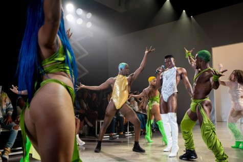 A group of models wearing green toned clothes dancing around a model wearing a white, see-through coverall and white boots holding a mic in the middle.