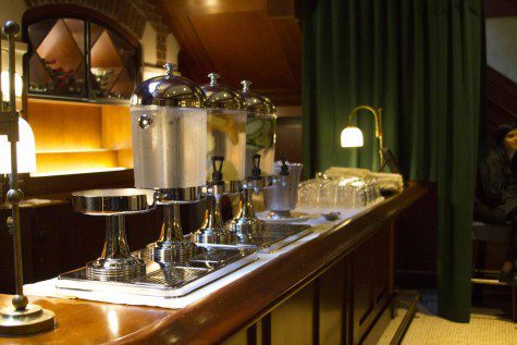 A diagonal view of a bar top with dispensers of water with lemons or cucumbers in the foreground and glass cups in the background.