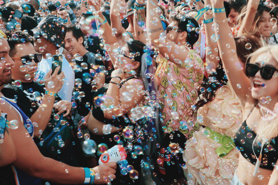 A close-up photograph of a crowd of concert attendees playing with bubbles. One attendee uses a bubble gun to make bubbles.